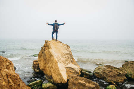 Traveler man with backpack standing on sandy beach in the middle of rocks against sea background. Place for text or advertising.の写真素材