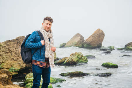 Traveler man with backpack standing on sandy beach in the middle of rocks against sea background. Place for text or advertising.の写真素材