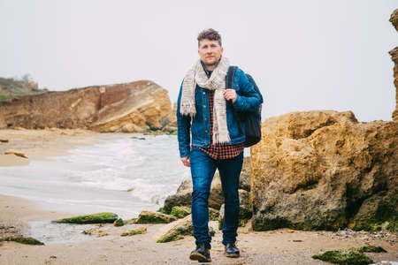 Traveler man with backpack standing on sandy beach in the middle of rocks against sea background. Place for text or advertising.の写真素材