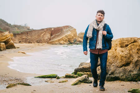 Traveler man with backpack standing on sandy beach in the middle of rocks against sea background. Place for text or advertising.の写真素材