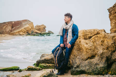 Traveler man with backpack standing on sandy beach in the middle of rocks against sea background. Place for text or advertising.の写真素材