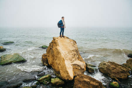 Traveler man with backpack standing on sandy beach in the middle of rocks against sea background. Place for text or advertising.の写真素材
