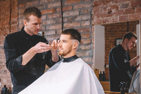 Young male barber shaves the client's head with a electric trimmer. Young bearded man getting haircut by hairdresser at barber shop. Advertising and barber shop concept.の写真素材