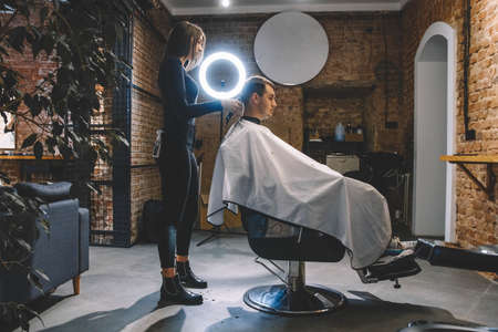Beautiful female hairdresser shaves the head of a client sitting in a chair with an electric trimmer in a barber shop. Concept of advertising and hairdressing.の写真素材
