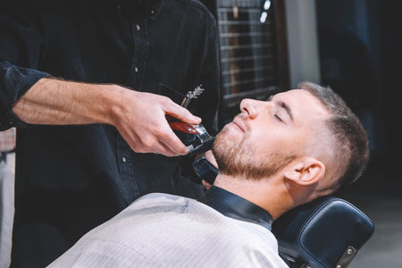 Young male barber shaves the client's head with a electric trimmer. Young bearded man getting...