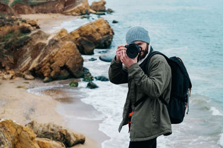 Young man photographer traveler with backpack taking pictures on sea and rocks background. Place for text or advertising.の写真素材