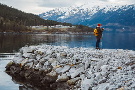 Traveler man with camera on tripod standing on the background of the mountain and a lake makes a photo landscape. Travel lifestyle concept. Copy, empty space for text.の写真素材