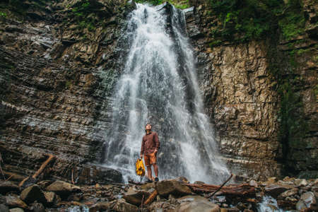 Traveler man with a yellow backpack standing on the background of a waterfall. Travel lifestyle conceptの写真素材