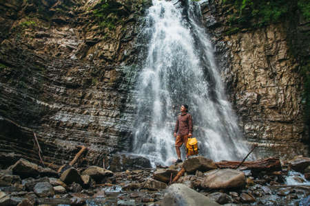 Traveler man with a yellow backpack standing on the background of a waterfall. Travel lifestyle conceptの写真素材