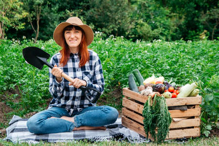 Beautiful woman farmer sitting on a blanket near fresh organic vegetables in a wooden box against the background of a vegetable garden. Copy, empty space for text.の写真素材
