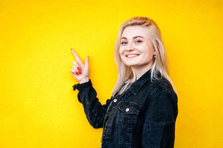 Beautiful blonde woman in black jacket smiling while lifting a finger up posing against a background of yellow wall. Copy, empty space for text.の写真素材