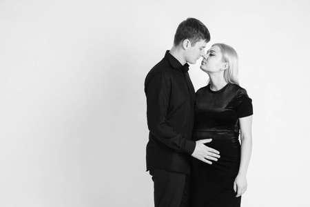 Pregnant woman and her husband hugging and holding onto your stomach in black clothes on a white background. Black and white picture. Beautiful married couple. Copy, empty space for text.の写真素材