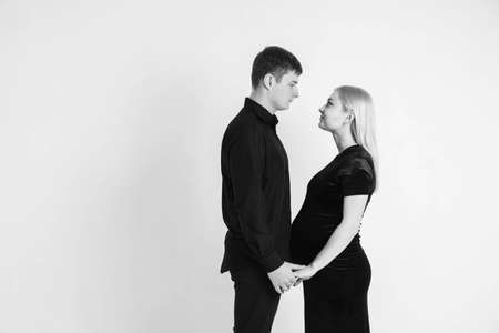Pregnant woman and her husband hugging and holding onto your stomach in black clothes on a white background. Black and white picture. Beautiful married couple. Copy, empty space for text.の写真素材