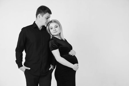 Pregnant woman and her husband hugging and holding onto your stomach in black clothes on a white background. Black and white picture. Beautiful married couple. Copy, empty space for text.の写真素材