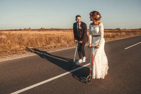 Smiling wedding couple riding a on scooters along the road outside the city at sunset. Place for text or advertising.の写真素材