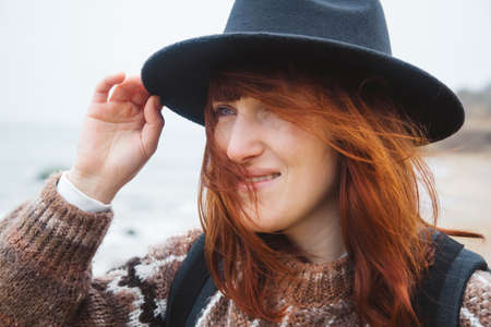 Beautiful red-haired woman in a hat and scarf with a backpack on the coast against the background of the rocks against the beautiful sea. Tourism, rest, lifestyle. Place for text or advertising.の写真素材