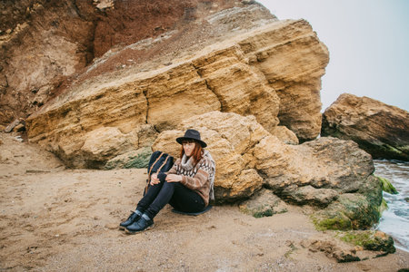 Beautiful red-haired woman in a hat and scarf with a backpack on the coast against the background of...