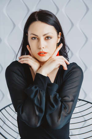 Beautiful woman in black dress posing near chair in bright studio. Place for text or advertising.の写真素材