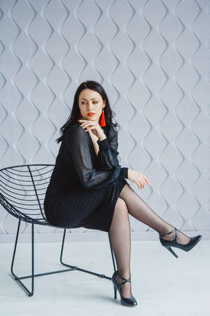 Beautiful woman in black dress posing near chair in bright studio. Place for text or advertising.の写真素材