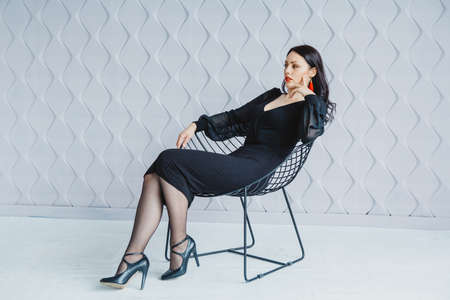 Beautiful woman in black dress posing near chair in bright studio. Place for text or advertising.の写真素材