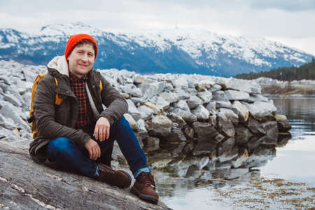 Young man with a yellow backpack wearing a red hat standing on a rock on the background of mountain and lake. Space for your text message or promotional content. Travel lifestyle concept.の写真素材