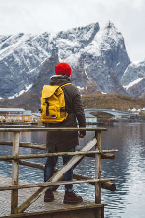 Traveler man is standing in the middle of a forest with a guitar on background of mountains and lake. Wearing a yellow backpack in a red hat. Place for text or advertising. Shoot from the back.の写真素材