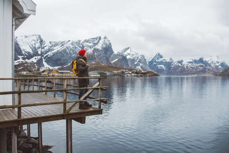 Traveler man is standing in the middle of a forest with a guitar on background of mountains and lake. Wearing a yellow backpack in a red hat. Place for text or advertising. Shoot from the back.の写真素材