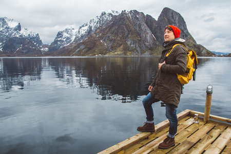 Traveler man is standing in the middle of a forest with a guitar on background of mountains and lake. Wearing a yellow backpack in a red hat. Place for text or advertising. Shoot from the back.の写真素材