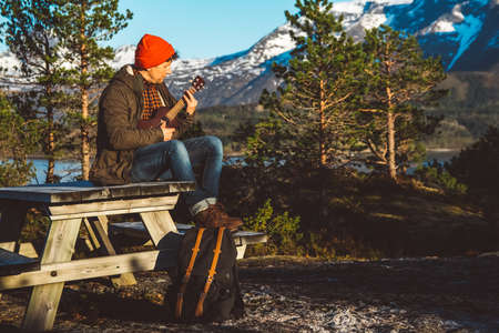 Traveler man is standing in the middle of a forest with a guitar on background of mountains and lake. Place for text or advertising. Shoot from the back.の写真素材