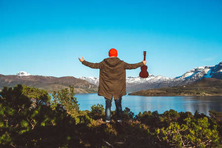 Traveler man is standing in the middle of a forest with a guitar on background of mountains and lake. Place for text or advertising. Shoot from the back.の写真素材