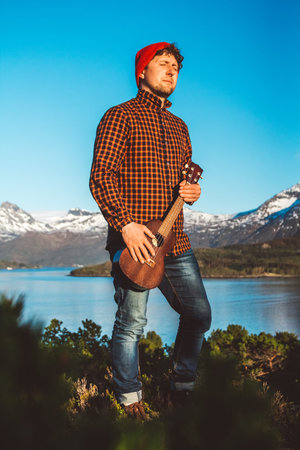 Young man is standing with a guitar on background of mountains and lake. Place for text or...