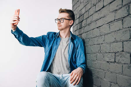 Portrait young man in shirt, jeans and glasses carrying a backpack and looking at smartphone while standing on a pink background. Copy, empty space for text.の写真素材