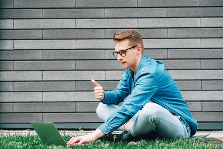 Young man in shirt and glasses he looks at the laptop in amazement and with a funny face sitting at the table on a white background. Funny face and amazement. Copy, empty space for text.の写真素材