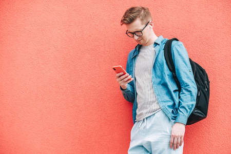 Young joyful man in shirt, jeans and glasses carrying a backpack and taking selfie photo on cellphone on a background of a pink wall. Copy, empty space for text.の写真素材