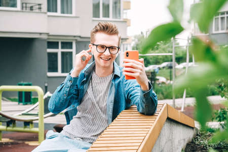 Young joyful man in shirt, jeans and glasses carrying a backpack and taking selfie photo on cellphone on a background of a pink wall. Copy, empty space for text.の写真素材