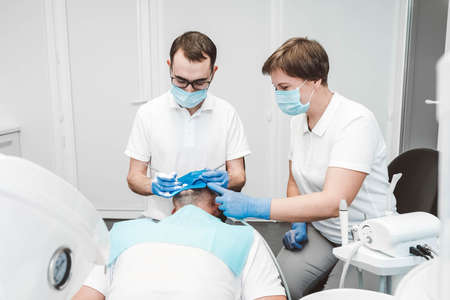 Dentist and his assistant treat a male patient in a dental clinic. Dentists in medical masks working with client's teeth. Copy, empty space for text.の写真素材