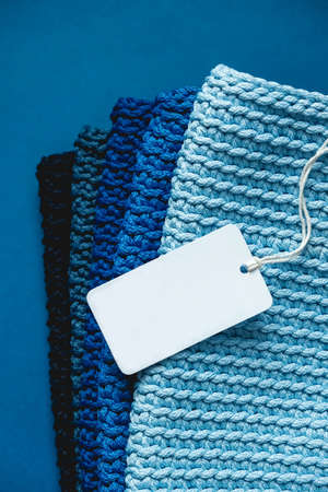 Stack of knitted material from threads of dark blue, light blue, gray colors with blank Price Tag on a blue background. Top view. Copy, empty space for text.の写真素材