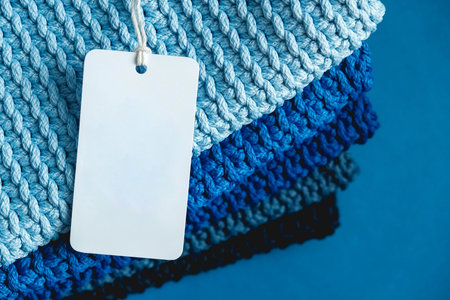 Stack of knitted material from threads of dark blue, light blue, gray colors with blank Price Tag on a blue background. Top view. Copy, empty space for text.の写真素材