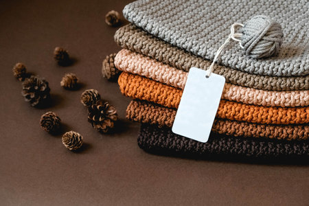 Stack of knitted material from threads of brown, orange, gray colors with an empty price tag on a brown background. Copy, empty space for text.の写真素材