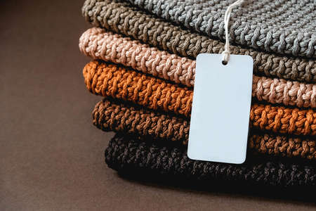 Stack of knitted material from threads of brown, orange, gray colors with an empty price tag on a brown background. Copy, empty space for text.の写真素材