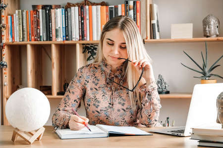 Beautiful woman wearing in blouse and glasses, sitting at the table with laptop and writing something in her notebook on the bookshelves background. Copy, empty space for text.の写真素材