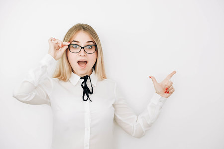 Pretty young blond woman wearing stylish white shirt and glasses over white background amazed and surprised pointing with finger at copy space. Copy, empty space for text.の写真素材