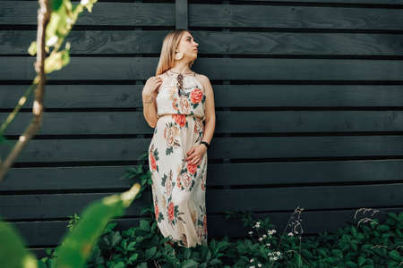 Beautiful blonde woman dressed in flower dress in a green leaves trees on a wooden wall background. Copy, empty space for text.の写真素材