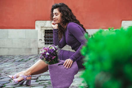 Young beautiful brunette woman wearing purple polka dot dress, holding knitted bag with flowers, posing in street of city. Copy, empty space for text.の写真素材