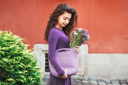 Young beautiful brunette woman wearing purple polka dot dress, holding knitted bag sitting on the stairs posing in street of city. Copy, empty space for text.の写真素材