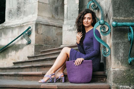 Young beautiful brunette woman wearing purple polka dot dress, holding knitted bag sitting on the stairs posing in street of city. Copy, empty space for text.の写真素材
