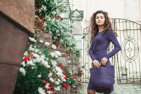 Young beautiful brunette woman wearing purple polka dot dress, holding knitted bag sitting on the stairs posing in street of city. Copy, empty space for text.の写真素材