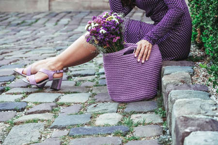 Young beautiful brunette woman wearing purple polka dot dress, holding knitted bag sitting on the stairs posing in street of city. Copy, empty space for text.の写真素材