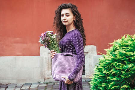 Young beautiful brunette woman wearing purple polka dot dress, holding knitted bag sitting on the stairs posing in street of city. Copy, empty space for text.の写真素材