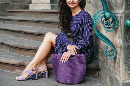 Young beautiful brunette woman wearing purple polka dot dress, holding knitted bag sitting on the stairs posing in street of city. Copy, empty space for text.の写真素材